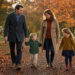 Une famille composée d’un homme, d’une femme et de deux enfants se promène dans un parc français aux couleurs de l’automne, baigné par une lumière dorée, avec une horloge de jardin discrète en arrière-plan.